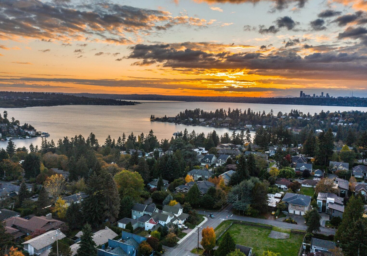 Aerial view of a suburban neighborhood with numerous trees, a lake, and a city skyline in the distance during sunset, with the sky filled with clouds.